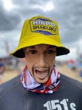 A mannequin or person-like display wearing a yellow bucket hat with a large yellow, blue, and white Hippo Suzuki logo patch on the front, plus a red, white, and blue neck gaiter, against a blurred outdoor British Superbikes BSB event background.