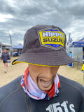 A mannequin or person-like display wearing a dark gray bucket hat with a large yellow, blue, and white Hippo Suzuki logo patch on the front, plus a red, white, and blue neck gaiter, against a blurred outdoor British Superbikes BSB event background.