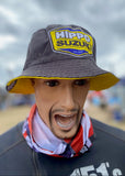 A mannequin or person-like display wearing a dark gray bucket hat with a large yellow, blue, and white Hippo Suzuki logo patch on the front, plus a red, white, and blue neck gaiter, against a blurred outdoor British Superbikes BSB event background.